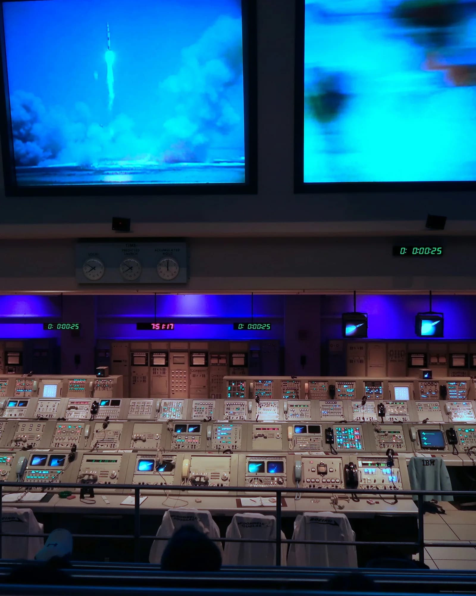 Mission-Control-Kontrollraum mit Konsolenreihen, Countdown-Uhren und Raketenstart auf der Leinwand